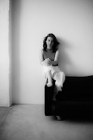 A person sits on the arm of a dark sofa against a plain, light-colored wall. The image is in black and white, creating a minimalist and introspective atmosphere. The person has an expressive, thoughtful gaze.