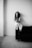 A person sits on the arm of a dark sofa against a plain, light-colored wall. The image is in black and white, creating a minimalist and introspective atmosphere. The person has an expressive, thoughtful gaze.