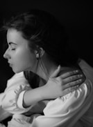 A black and white portrait of a young woman with closed eyes, her long hair partially tied back, wearing a light-colored garment. She rests her left hand on her right shoulder, conveying a serene and contemplative demeanor.