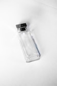 A clear, rectangular glass bottle with a metallic square cap. The bottle appears empty and is photographed against a plain white background, giving a minimalist aesthetic.