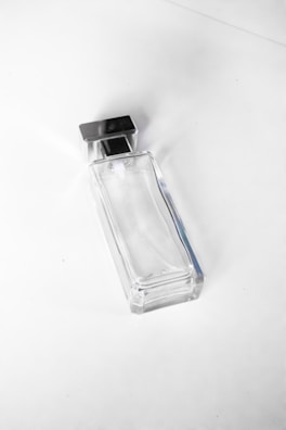 A clear, rectangular glass bottle with a metallic square cap. The bottle appears empty and is photographed against a plain white background, giving a minimalist aesthetic.