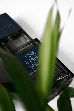 A bottle of perfume labeled 'One Man Show' is partially obscured by green leaves. The bottle has a dark cap and the text on the bottle is visible. The focus is slightly softened, giving a natural and refreshing look.