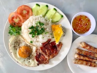 A well-arranged plate of Vietnamese-style rice is accompanied by a fried egg, sliced vegetables including tomatoes and cucumbers, grilled pork with black pepper, and fresh herbs. A side of Vietnamese egg meatloaf is present along with a small bowl of dipping sauce containing chili and sesame seeds. Three grilled sausages are placed on a separate plate.