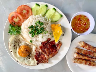 A well-arranged plate of Vietnamese-style rice is accompanied by a fried egg, sliced vegetables including tomatoes and cucumbers, grilled pork with black pepper, and fresh herbs. A side of Vietnamese egg meatloaf is present along with a small bowl of dipping sauce containing chili and sesame seeds. Three grilled sausages are placed on a separate plate.