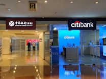 A hallway in a shopping mall features two bank outlets side by side: Bank of China (Hong Kong) on the left and Citibank on the right. Both banks have illuminated signs with their logos. The Bank of China section shows a closed office area with some ATMs in use by customers. The Citibank area has a bright, blue-lit interior with a welcoming entrance.