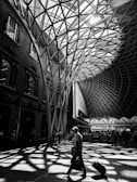 A striking architectural space featuring an intricate web-like roof structure allows sunlight to cast dynamic shadows across the floor. A lone traveler with a suitcase moves through the space, while other individuals are visible in the background near the building's walls and entrances.