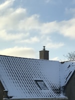 A snow-covered roof with a brick chimney against a clear blue sky. There is a small window visible on the roof and a few bare tree branches in the background.