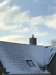 A snow-covered roof with a brick chimney against a clear blue sky. There is a small window visible on the roof and a few bare tree branches in the background.