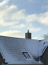 A snow-covered roof with a brick chimney against a clear blue sky. There is a small window visible on the roof and a few bare tree branches in the background.