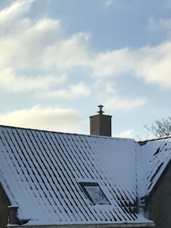 A snow-covered roof with a brick chimney against a clear blue sky. There is a small window visible on the roof and a few bare tree branches in the background.