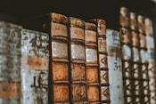 Several antique books are lined up in a row on a shelf. The books have worn and aged covers, with some displaying Roman numerals on their spines. The bindings appear to be made of leather and show signs of significant wear and patina, indicating their age.