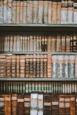 An assortment of vintage books arranged on wooden shelves, exhibiting leather-bound covers with rich textures and embossed patterns. The books vary in color, featuring shades of brown, beige, and muted greens, with titles and labels visible on the spines.