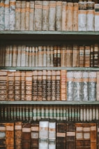 An assortment of vintage books arranged on wooden shelves, exhibiting leather-bound covers with rich textures and embossed patterns. The books vary in color, featuring shades of brown, beige, and muted greens, with titles and labels visible on the spines.