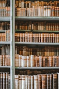 A collection of vintage books lined up on wooden shelves, showcasing a variety of worn spines with different colors and patterns. The books appear to be arranged by size and the shelves are tall, reaching nearly from floor to ceiling, giving a sense of an old library or study. The image has a slightly aged, sepia tone, enhancing the historic feel.
