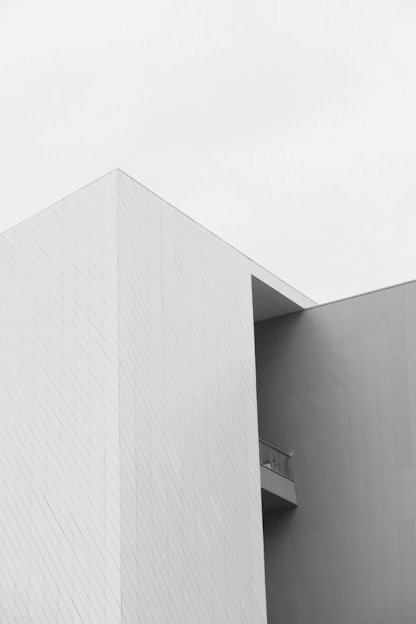 A minimalist architectural structure with clean lines and a geometric shape is depicted. The building has a stark, white facade with subtle linear patterns. A small balcony with a glass railing is visible, adding a touch of detail to the otherwise simple design.