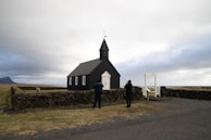 A small black church with a pointed steeple stands on an open landscape. Two people are taking photos near the church. The sky is overcast, and there are stone walls surrounding parts of the area. In the background, there is a view of the sea and distant mountains.