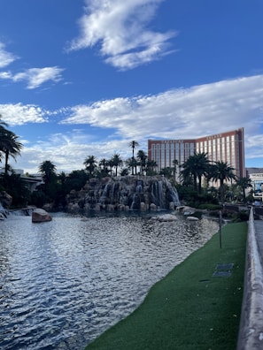 A scenic view of a man-made waterfall surrounded by palm trees, with a large hotel building in the background under a vibrant blue sky filled with clouds.