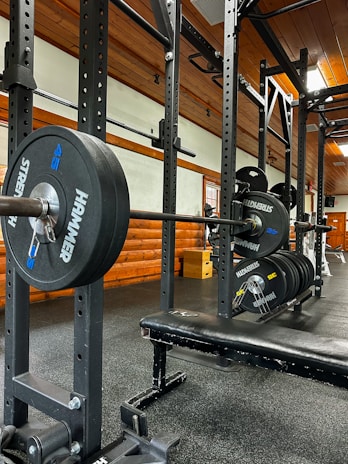 A gym setting featuring a squat rack with a barbell loaded with several weight plates, prominently branded with 'HAMMER' and 'STRENGTH'. The gym has wooden walls and ceiling, providing a rustic look. A leather bench press seat is positioned in front of the squat rack. Various weightlifting equipment and a window with white frames are visible.
