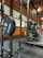 A gym setting featuring a squat rack with a barbell loaded with several weight plates, prominently branded with 'HAMMER' and 'STRENGTH'. The gym has wooden walls and ceiling, providing a rustic look. A leather bench press seat is positioned in front of the squat rack. Various weightlifting equipment and a window with white frames are visible.