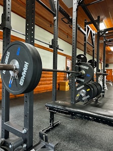 A gym setting featuring a squat rack with a barbell loaded with several weight plates, prominently branded with 'HAMMER' and 'STRENGTH'. The gym has wooden walls and ceiling, providing a rustic look. A leather bench press seat is positioned in front of the squat rack. Various weightlifting equipment and a window with white frames are visible.