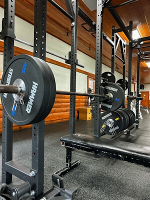 A gym setting featuring a squat rack with a barbell loaded with several weight plates, prominently branded with 'HAMMER' and 'STRENGTH'. The gym has wooden walls and ceiling, providing a rustic look. A leather bench press seat is positioned in front of the squat rack. Various weightlifting equipment and a window with white frames are visible.