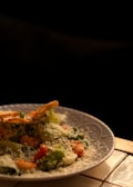 A plate of salad is garnished with grated cheese, featuring a mix of fresh greens, sliced tomatoes, and possibly croutons or crispy toppings. The lighting creates a warm and inviting atmosphere, highlighting the textures and ingredients.
