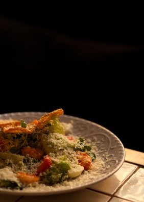 A plate of salad is garnished with grated cheese, featuring a mix of fresh greens, sliced tomatoes, and possibly croutons or crispy toppings. The lighting creates a warm and inviting atmosphere, highlighting the textures and ingredients.