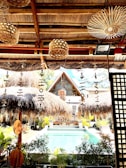 A tropical setting featuring a thatched-roof structure overlooking a swimming pool. The foreground includes rustic decorations such as wooden wind chimes and natural woven lampshades. Lush greenery surrounds the pool area, with a bohemian aesthetic complemented by natural materials.