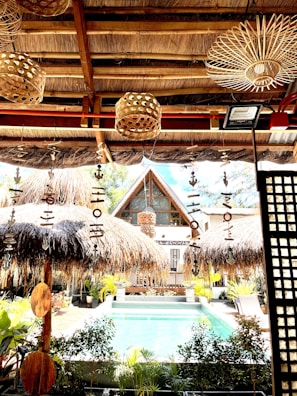 A tropical setting featuring a thatched-roof structure overlooking a swimming pool. The foreground includes rustic decorations such as wooden wind chimes and natural woven lampshades. Lush greenery surrounds the pool area, with a bohemian aesthetic complemented by natural materials.