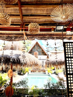 A tropical setting featuring a thatched-roof structure overlooking a swimming pool. The foreground includes rustic decorations such as wooden wind chimes and natural woven lampshades. Lush greenery surrounds the pool area, with a bohemian aesthetic complemented by natural materials.