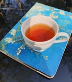 A white cup filled with tea is placed on a notebook with a blue cover featuring floral designs. The scene is outdoors, with a reflective surface underneath showing blurry reflections of trees.