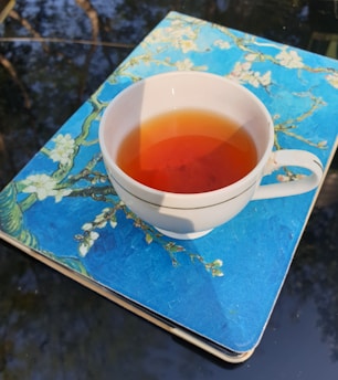 A white cup filled with tea is placed on a notebook with a blue cover featuring floral designs. The scene is outdoors, with a reflective surface underneath showing blurry reflections of trees.