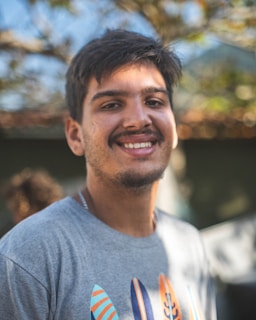 A young man with dark hair and a mustache is smiling. He is wearing a gray t-shirt with colorful surfboard designs. The background is softly blurred, with hints of green and brown, suggesting an outdoor setting.