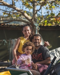A joyful family portrait with a smiling man, woman, and child in an outdoor setting. The man and woman are sitting close together, while the child is in the foreground, making a playful gesture. The background features a tree with green and yellow leaves, suggesting a pleasant sunny day.