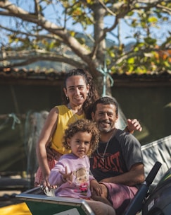 A joyful family portrait with a smiling man, woman, and child in an outdoor setting. The man and woman are sitting close together, while the child is in the foreground, making a playful gesture. The background features a tree with green and yellow leaves, suggesting a pleasant sunny day.
