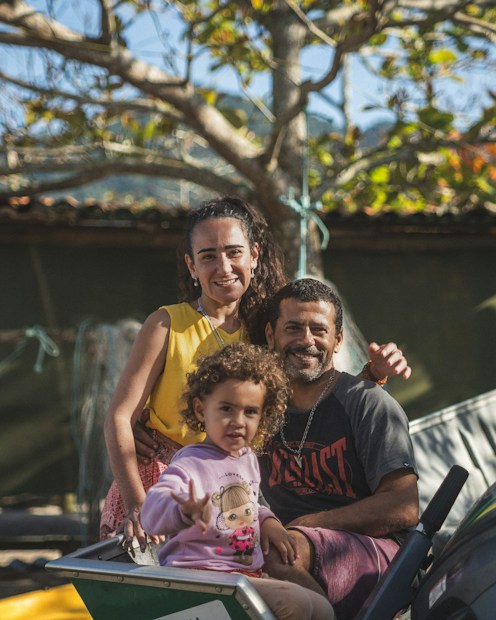 A joyful family portrait with a smiling man, woman, and child in an outdoor setting. The man and woman are sitting close together, while the child is in the foreground, making a playful gesture. The background features a tree with green and yellow leaves, suggesting a pleasant sunny day.