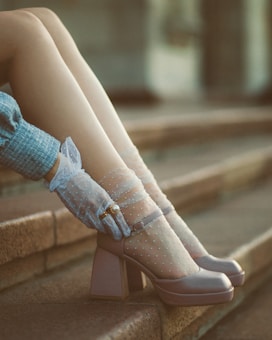A pair of legs in sheer dotted socks and stylish high-heeled shoes are elegantly displayed on stone steps. The wearer also dons delicate gloves, adorned with a pearl ring, which adds a touch of vintage charm to the ensemble.