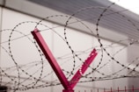 Barbed wire spirals tightly wound around a red metal post, positioned against a backdrop of a muted gray building. The wire exhibits a repeating pattern with sharp barbs, and the scene conveys a sense of restriction or security.