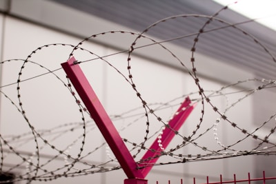 Barbed wire spirals tightly wound around a red metal post, positioned against a backdrop of a muted gray building. The wire exhibits a repeating pattern with sharp barbs, and the scene conveys a sense of restriction or security.