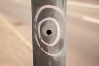 A cylindrical metal pole with a small circular hole in the center. It has white and pink spray-painted circular patterns around the hole. The background is blurred, showing part of a pavement and a road.