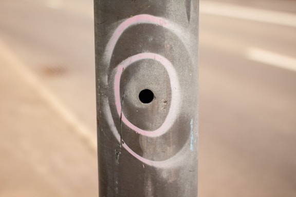A cylindrical metal pole with a small circular hole in the center. It has white and pink spray-painted circular patterns around the hole. The background is blurred, showing part of a pavement and a road.