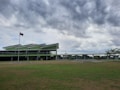 A modern green building with a distinctive roof design set against a backdrop of cloudy skies. It is situated in a grassy area with a flagpole displaying the Philippine flag.