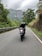 A motorcycle with two riders is traveling down a winding, tree-lined road. The road is bordered by lush green foliage and a rocky slope, with distant hills visible under an overcast sky.