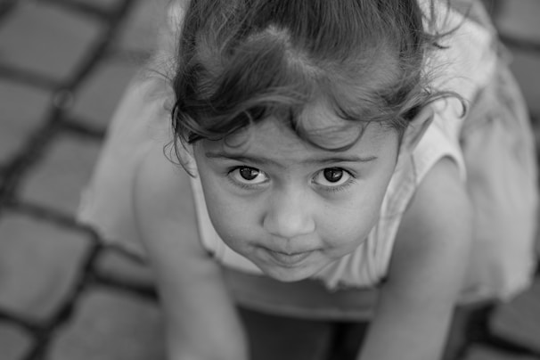 A young child with curly hair is looking directly into the camera with large, expressive eyes. The child appears to be kneeling or crouching on a cobblestone surface. The image is in black and white, emphasizing the texture of the child's hair and the surface beneath.