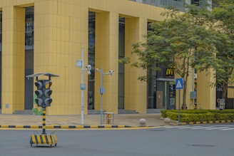 A street scene in an urban area features a modern building with large yellow vertical panels. In the foreground, there is a portable traffic signal system on wheels, surrounded by various surveillance cameras. A McDonald's restaurant is visible in the background, with its entrance framed by greenery and trees. The street is marked with crossing lines and bordered by curbs with yellow and black stripes.