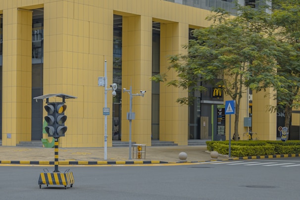 A street scene in an urban area features a modern building with large yellow vertical panels. In the foreground, there is a portable traffic signal system on wheels, surrounded by various surveillance cameras. A McDonald's restaurant is visible in the background, with its entrance framed by greenery and trees. The street is marked with crossing lines and bordered by curbs with yellow and black stripes.
