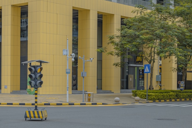 A street scene in an urban area features a modern building with large yellow vertical panels. In the foreground, there is a portable traffic signal system on wheels, surrounded by various surveillance cameras. A McDonald's restaurant is visible in the background, with its entrance framed by greenery and trees. The street is marked with crossing lines and bordered by curbs with yellow and black stripes.