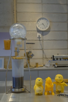 A laboratory setup with a cold drip coffee maker on a metal table. Three small yellow toy figures are positioned next to the coffee maker. The background consists of a clock on a cement wall, some machinery, and countertop items, creating a blend of industrial and playful elements.