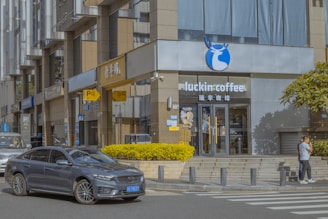 A storefront for a coffee shop named Luckin Coffee, with a gray car parked on the street in front. The building exterior is modern, featuring large windows and a blue and white company logo. There is a yellow hedge lining the sidewalk, and two people are walking nearby. Various signs and banners are visible on adjacent buildings.