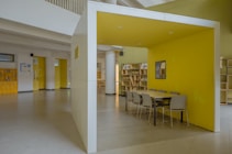 A modern indoor space featuring a yellow-walled booth with a long table and several gray chairs. The area is well-lit and spacious, with minimalistic design elements. Bookshelves line the walls, and there are various doors leading to adjacent rooms.